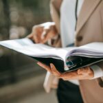 Crop faceless female entrepreneur in formal jacket turning pages of documents in folder while reading